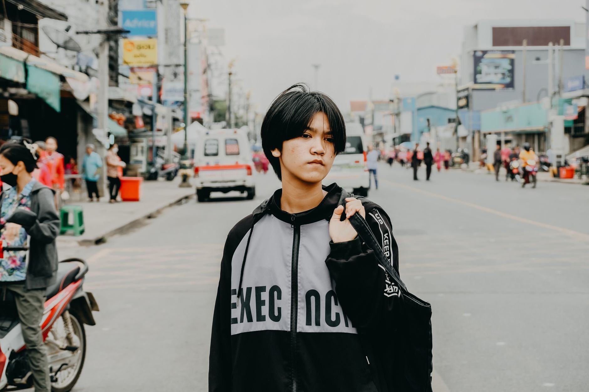 A young person with a jacket stands confidently in an urban street environment.