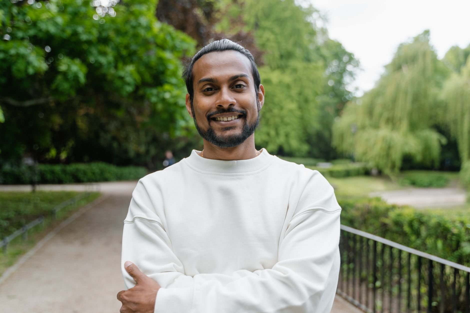 Smiling man in a white sweater stands confidently in a green park, embodying positivity and relaxation.